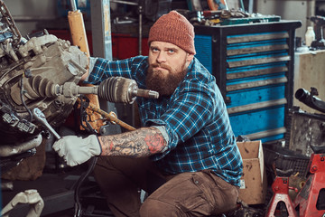A brutal tattooed bearded mechanic specialist repairs the car engine which is raised on the hydraulic lift in the garage. Service station.