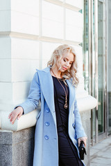 human emotion. Urban style. Portrait of a cute young blonde in a fashionable blue coat. The girl posing against the backdrop of urban architecture.