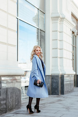 human emotion. Urban style. Portrait of a cute young blonde in a fashionable blue coat. The girl posing against the backdrop of urban architecture.