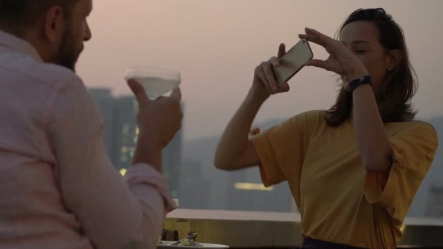 Woman taking photo of her boyfriend with cocktail sitting on terrace in bar
