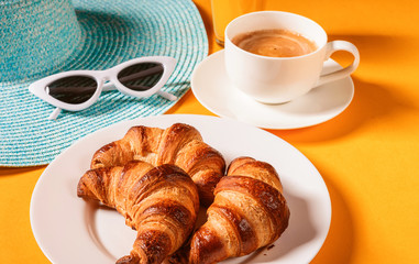 hat, sunglasses croissant with a cup of coffee and a glass of orange juice on a yellow table in the sun
