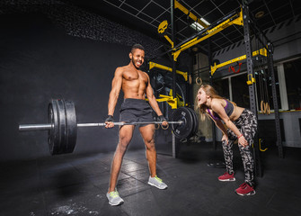 fit woman girl motivates african man while he doing deadlift with heavy barbell. man lifting barbell opposite window. emotional moment of lifting weight. affective lifting in gym. sport couple in gym.