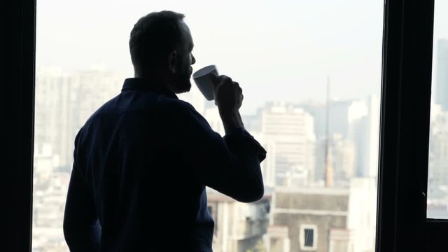 Silhouette of man with beverage admire view from window at home

