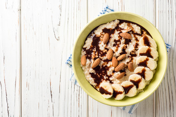 Oatmeal porridge with banana, almonds and chocolate syrup on white wooden background