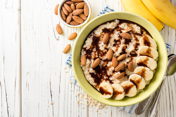 Oatmeal porridge with banana, almonds and chocolate syrup on white wooden background