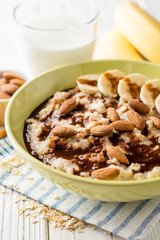 Oatmeal porridge with banana, almonds and chocolate syrup on white wooden background