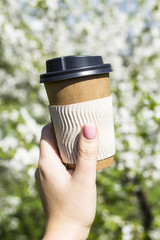 A woman's hand holds a disposable cup of coffee against the blossoming spring tree. The beginning of a beautiful spring day. Good mood and lifestyle