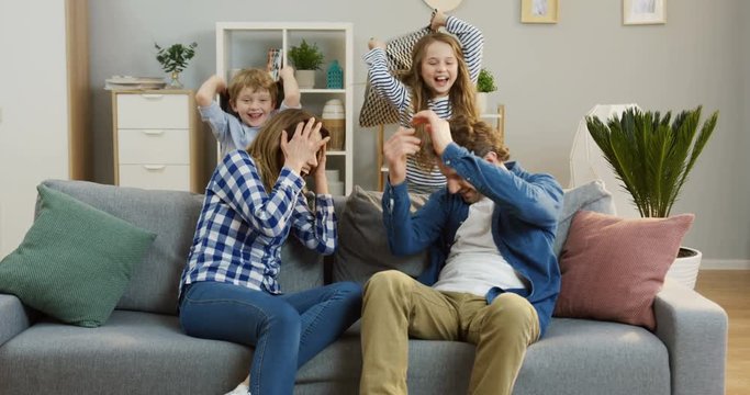 Young parents sitting on the sofa in the living room and their funny cheerful kids beating them with pillows. Family having fun. Indoor