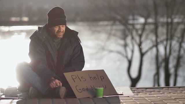 Lonely homeless sitting near river bank begging for money with handwritten sign. Woman passing by and giving bearded mature male a cup of hot coffee. Beggar drinking coffee and smiling