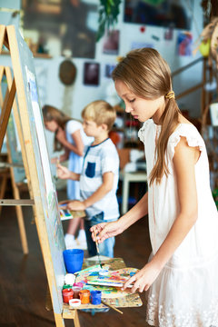Side view portrait of pretty girl painting picture on easel in art class with other children in background