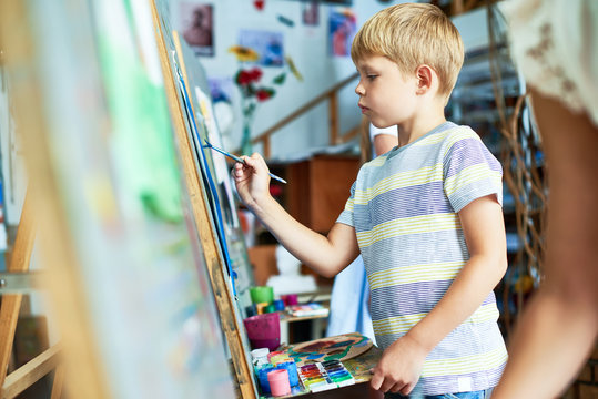 Side view portrait of cute  boy painting on easel during art class, concentrating on his masterpiece