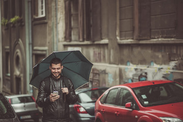 Young happy man using his cell phone, walking the streets on a rainy day and holding an umbrella