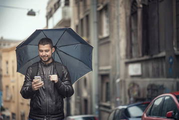 Young happy man using his cell phone, walking the streets on a rainy day and holding an umbrella