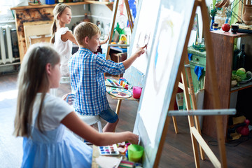 Side view portrait of three children painting on easels during art class in sunlit studio, focus on talented little boy in the middle