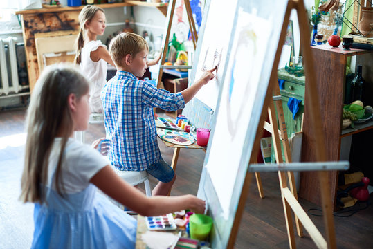 Side view portrait of three children painting on easels during art class in sunlit studio, focus on talented little boy in the middle