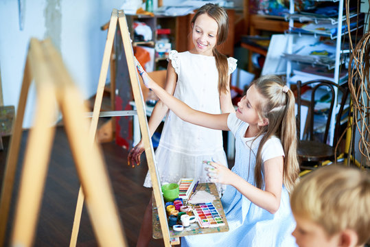 Portrait of pretty girl painting picture on easel in art class, with other children watching her, copy space