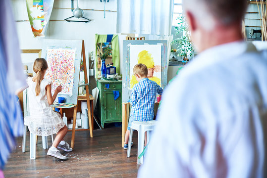 Back view portrait of senior teacher watching children painting on easels during art class  in cozy studio decorated with plants