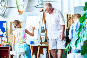 Portrait of senior art teacher leaning over easel to watch little student painting pictures on easel in art school, copy space