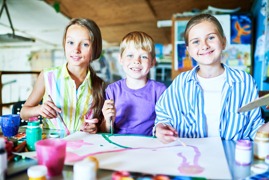 Portrait of three cheerful little kids painting pictures in art class sitting at tables with art supplies, pencils and paints, and looking at camera smiling happily
