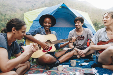 Group of young adult friends in camp site playing guitar and ukelele and singing together outdoors...