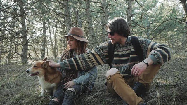 Young couple with backpacks in spring forest, hikers with corgi dog relaxing in sunny forest 