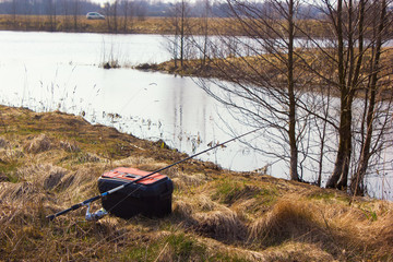 Fishing spnning rod and box with fishing gear standing on the river bank. Around are young trees. In the background, in the distance, there is a car on the other side. Sunny spring day.