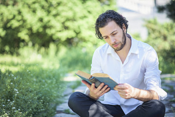 Fototapeta premium Young intellectual man reading a book in the park, enjoying his hobby all by himself, surrounded by tall green grass