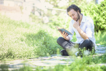 Young intellectual man reading a book in the park, enjoying his hobby all by himself, surrounded by tall green grass