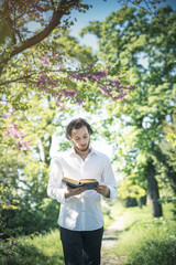 Young intellectual man reading a book in the park, enjoying his hobby all by himself, surrounded by tall green grass