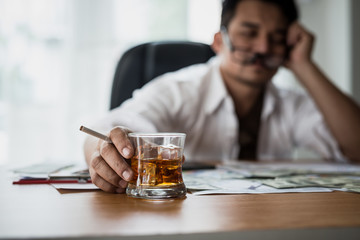Stressed asian businessman holding a glass of whiskey and smoking. he sleep on the money, data charts, business document at office desk.  alcohol addiction - drunk businessman concept.