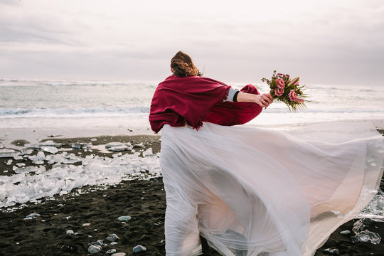 Bride in white flattering dress with a bouquet walking in windy day on Black Sand Beach, Iceland. Artwork. Her hair shakes by wind