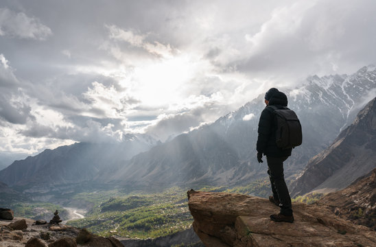 a man with backpack standing on mountain peak and bright sunlight through cloudy sky. Travel lifestyle and achievement success concept