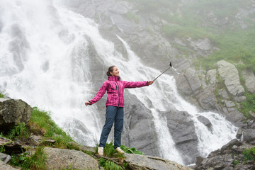 Pretty young woman taking a selfie in front of big powerful waterfall Balea in Romanian Fagarash mountains. Beautiful smiling girl travel in nature. Traveling and recreation concept.