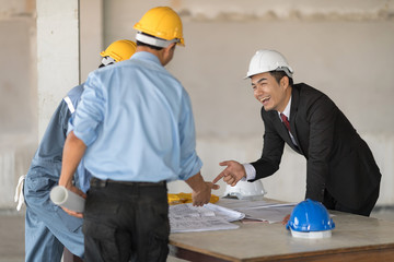 Businessman in white helmet with black suit and red necktie is look at architect and pointing at blueprint. He is laughing. Blur foreground are engineer and architect in the yellow helmet.