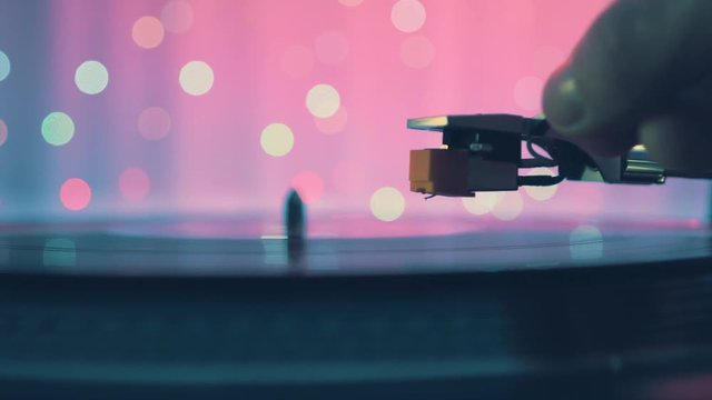The moment of a rotating vinyl plate turntable on a pink background and bright bokeh lights. Sound technology for DJ to mix and play music. The man's hand puts and removes needle from the vinyl plate 