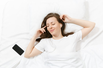 Top view of happy young woman lying in bed with white sheet, pillow, blanket, listen music from earphones in mobile phone. Calm beautiful female spending time in room. Rest, relax, good mood concept.