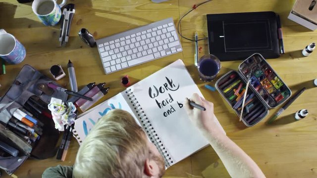 Top view of young male illustrator using black watercolor paint to write beautiful text saying Work Hard and Be Nice, in his sketchbook