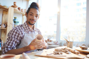 Portrait of young mixed race man holding small clay vase in workshop and looking at camera with satisfied smile