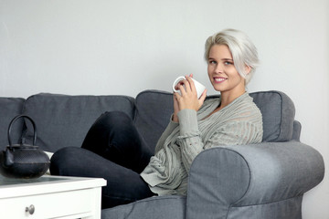 Hübsche blonde Frau sitzt lächelnd auf einer Couch und hält eine Tasse in ihren Händen 