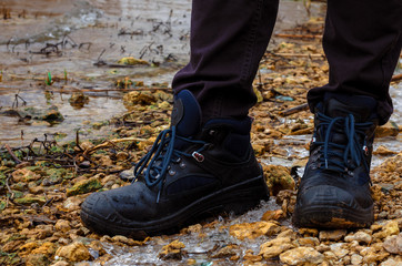 A lonely man in dark jeans and heavy road boots on lacing strolls in the cold season stands on the  river bank. On the shore, small stones, ice, thickets of reeds and shrubs.