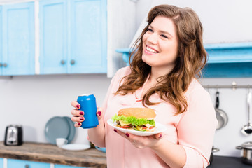 overweight smiling woman with soda drink and burger in hands in kitchen at home