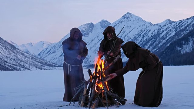 Group of monks in brown cloaks of the order of St. Francis are warming themselves by the fire