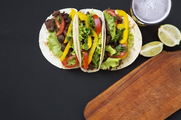 Tacos with meat and vegetables, beer and lime on a black wooden background, top view. Black background for text and design. Flat top view.