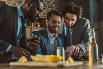 smiling multiethnic men using smartphone and drinking whisky together