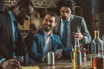 cheerful multiethnic men in suits using smartphone and drinking alcoholic beverages together