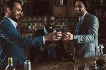 cheerful young male friends in suits clinking glasses of whiskey