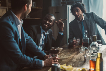 smiling multiethnic men talking, drinking whiskey and smoking cigars while partying together