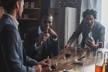 three multiethnic men in suits smoking cigars and drinking alcohol together - obrazy, fototapety, plakaty