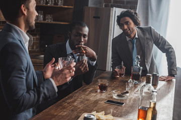cropped shot of smiling multiethnic businessmen talking while smoking cigars and drinking whiskey