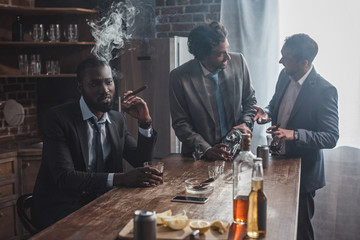 three multiethnic men in suits drinking alcohol beverages and smoking cigars together